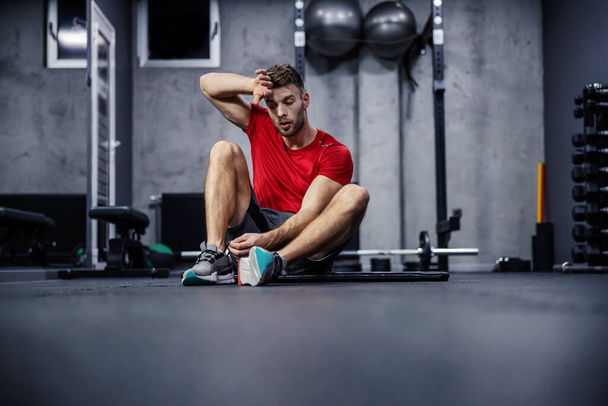 Усталость в тренировках. Мужчина сидит на полу спортзала и отдыхает между упражнениями. Он устал и вытирает пот с лица рукой. Устойчивость и потеря веса - Фото, изображение