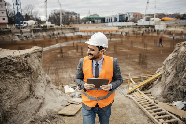 Ein gut gelaunter Bauunternehmer überprüft die Arbeiten auf der Baustelle und hält ein Tablet in der Hand. - Foto, Bild