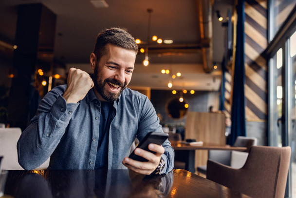 Un giovane uomo di successo sta celebrando la vittoria in caffetteria mentre guarda il suo telefono. - Foto, immagini