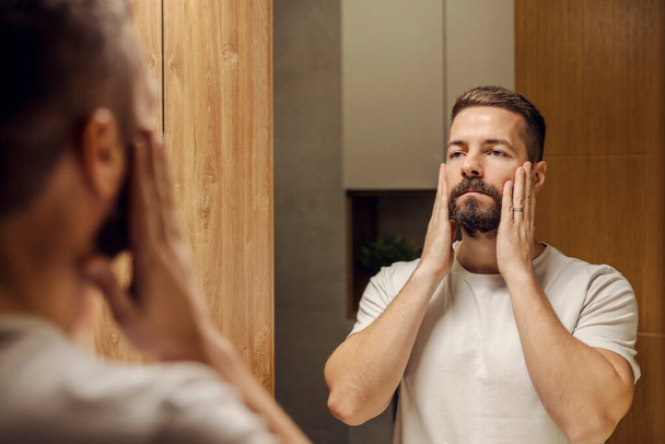 Een man met een baard raakt zijn gezicht aan en kijkt naar zijn rimpels in de spiegel in de badkamer. - Foto, afbeelding