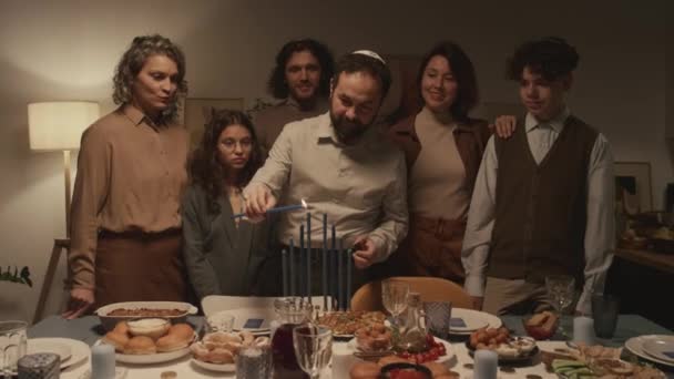 Tiro médio da família judaica de seis em torno da mesa de jantar na celebração Hanukkah, homem em kippah segurando champô ardente e acendendo velas em hanukkiah, enquanto todo mundo está assistindo - Filmagem, Vídeo
