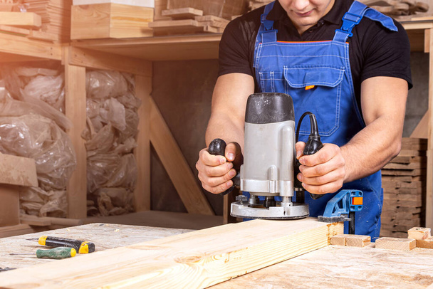  Un joven carpintero moreno equivale a un tablón de madera con una fresadora en el taller - Foto, Imagen