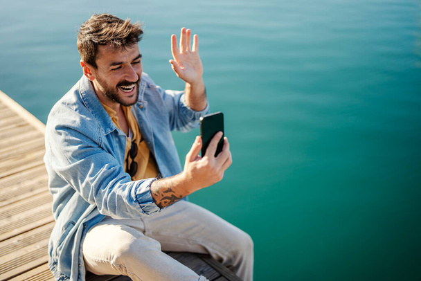 Egy boldog ember üdvözli a barátait, miközben a kikötőben ül, és videóhívást tart a telefonján.. - Fotó, kép