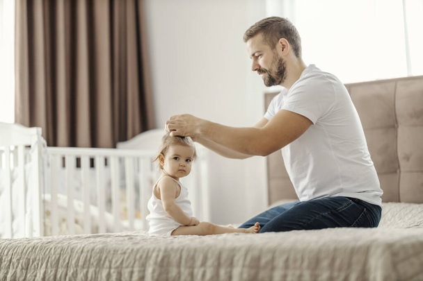 Un padre está haciendo el pelo de su hija mientras está sentado en la cama en casa. - Foto, Imagen