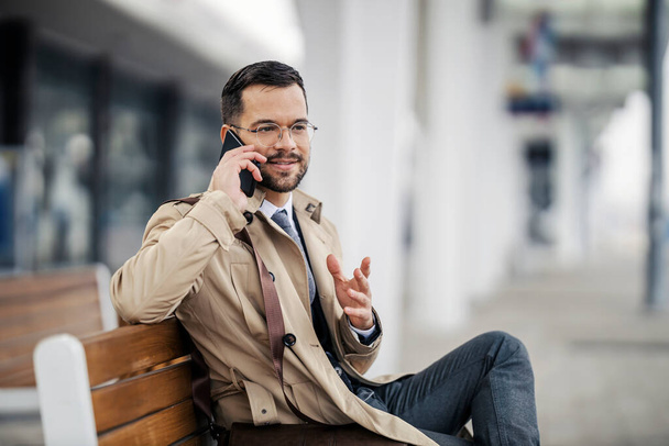 A happy young smart casual businessman is sitting on a bench at the station and waiting for a train while talking on the phone. - Photo, Image