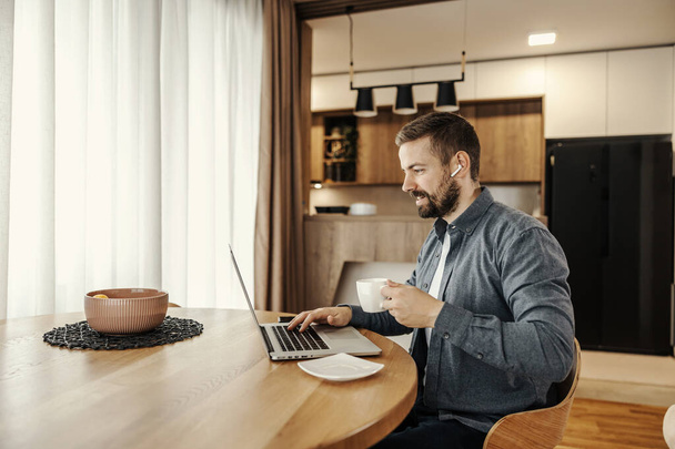 Un allegro lavoratore remoto si sta godendo il suo caffè mentre lavora da casa su un computer portatile. - Foto, immagini