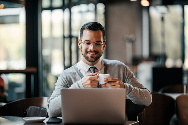 Un giovane uomo d'affari informale intelligente sta bevendo caffè in mensa mentre sorride al computer portatile. - Foto, immagini