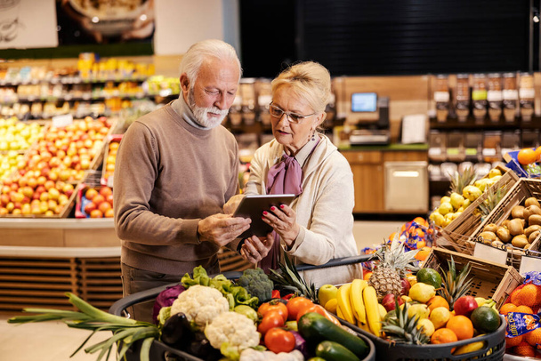A senior couple is looking and following groceries list on tablet at the supermarket. - Foto, imagen