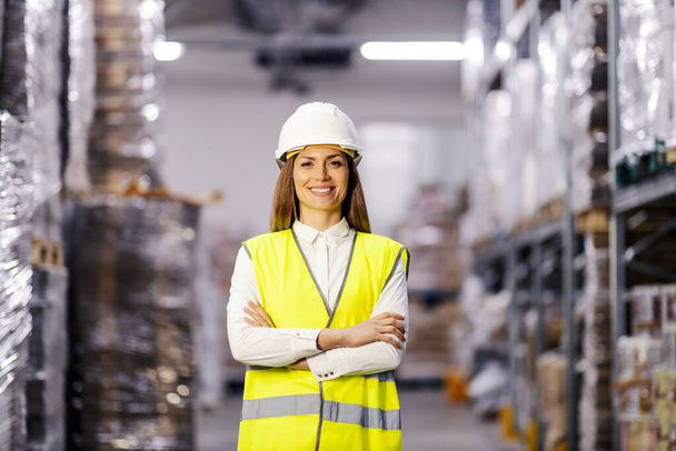Portrait of a confident female manager standing in storage with arms crossed and smiling at the camera. - Фото, зображення