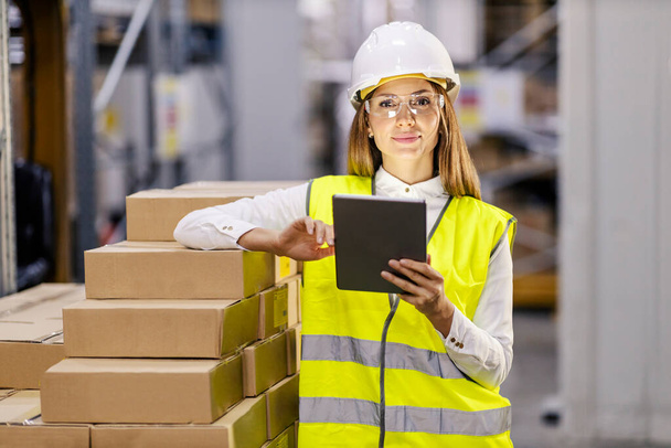 An inventory manager in work wear is using tablet in warehouse and looking at the camera. - 写真・画像