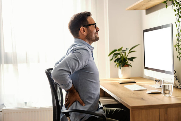 Un freelancer se sienta en su silla en la oficina en casa y tiene dolor de espalda. - Foto, imagen