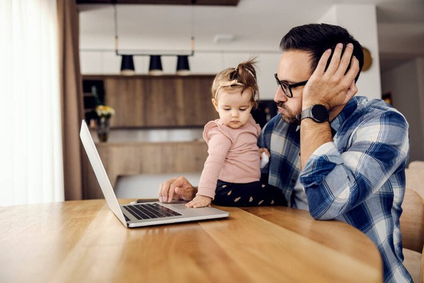Un padre impegnato cerca di finire il lavoro su un computer portatile e la sua bambina lo sconvolge mentre è seduto a casa. - Foto, immagini