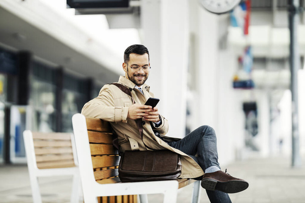 Een jonge elegante zakenman zit op een bankje op het station en wacht op een transport terwijl hij glimlacht aan de telefoon. - Foto, afbeelding