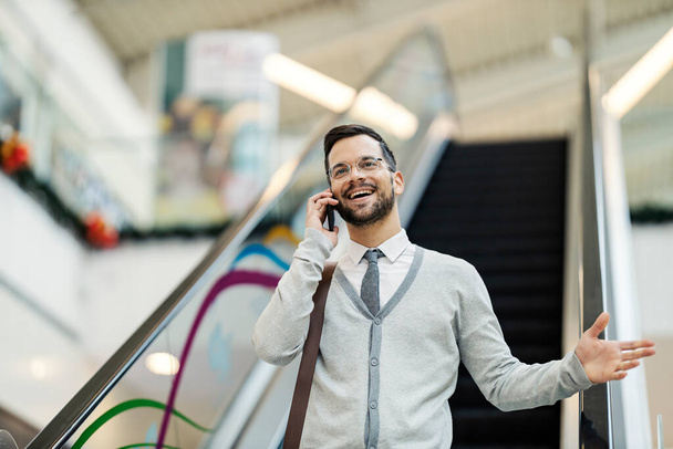 Un joven feliz hombre en casual inteligente en escaleras mecánicas en el centro comercial está hablando por teléfono. - Foto, Imagen