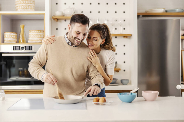 Щасливий чоловік перемішує яйця на сковороді вдома, а його дружина обіймає його. - Фото, зображення