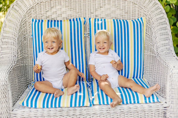 Gémeos doces, uma família cheia de amor. Os bebês bonitos nos bodysuits brancos gostam de comer biscoitos e enquanto se sentam em uma grande cadeira de lona com almofadas coloridas. Gêmeos com olhos azuis e cabelo loiro - Foto, Imagem