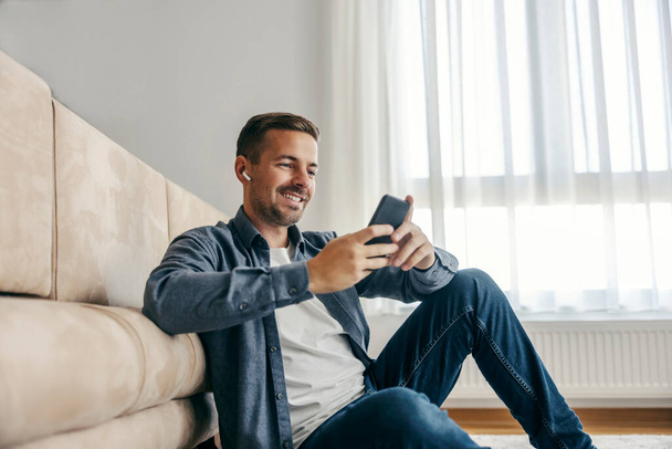 Egy boldog férfi ül otthon a padlón és mosolyog a telefonra.. - Fotó, kép