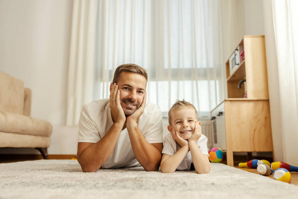 Portret szczęśliwego taty i dziecka leżących na podłodze w domu i uśmiechających się do kamery. - Zdjęcie, obraz