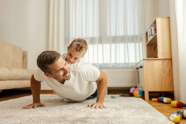 Un hijo en la espalda del padre lo está mirando mientras papá está haciendo flexiones en casa y practicando. - Foto, imagen