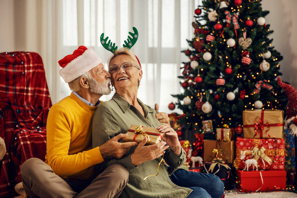 Старший чоловік дарує подарунок дружині, цілує і обіймає її на Різдво і Новий рік вдома.. - Фото, зображення