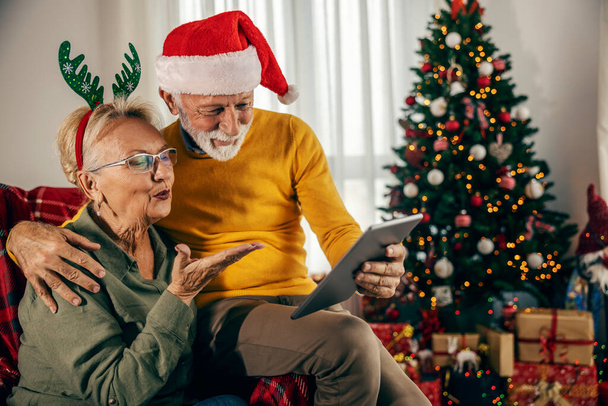 Una pareja mayor que tiene videollamada en la tableta en casa con la familia y el envío de besos en Navidad y año nuevo. - Foto, imagen