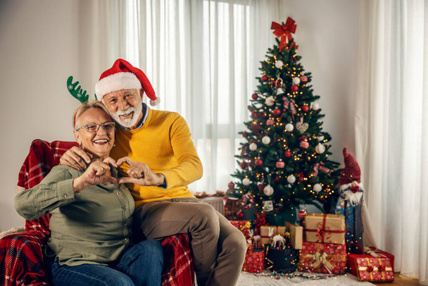 Счастливая пожилая пара делает жест в форме сердца, обнимаясь и сидя дома на Рождество и Новый год. - Фото, изображение