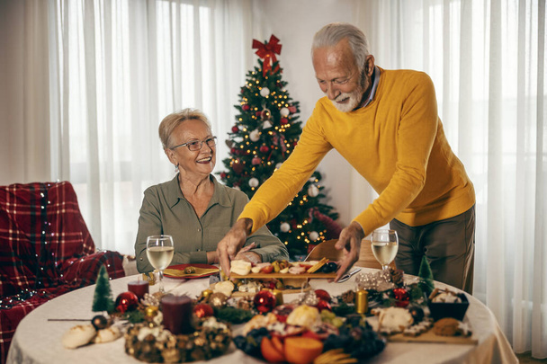 Ein älterer Mann bringt an Weihnachten und Neujahr zu Hause Essen auf den Weihnachtstisch. - Foto, Bild