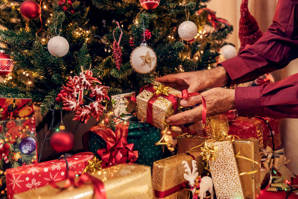 クリスマスと新年のクリスマスツリーの下にクリスマスプレゼントを置く上級男性の手の近く. - 写真・画像