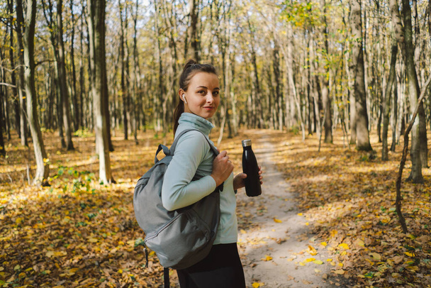 The girl drinks water from an iron bottle, not plastic. A beautiful girl is doing fitness outdoors in a sunny autumn forest. Body positive, sports for women, healthy lifestyle, self-love and wellness. - Photo, Image
