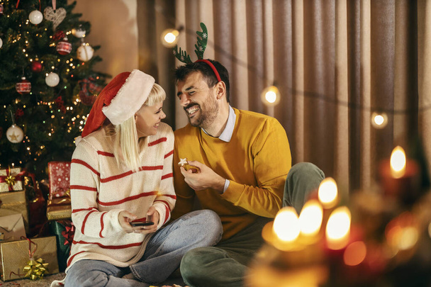 Ένα χαρούμενο νεαρό ζευγάρι κάθεται στο σπίτι κοντά στο χριστουγεννιάτικο δέντρο και γιορτάζει τα Χριστούγεννα και την παραμονή της Πρωτοχρονιάς. - Φωτογραφία, εικόνα