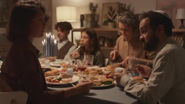 Közepes lövés zsidó család ül körül ünnepi asztal és élvezi a hagyományos ételek Hanuka vacsora, apa kippah beszél női rokon vagy barát, majd mindketten nevetnek - Felvétel, videó