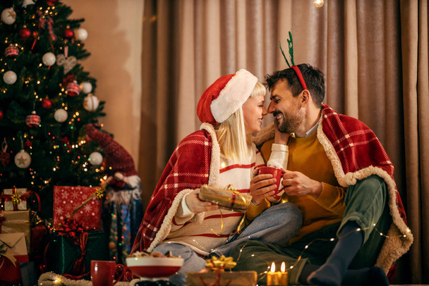 Una feliz pareja está sentada en casa cerca del árbol de Navidad, abrazando e intercambiando regalos en Navidad y víspera de año nuevo. - Foto, imagen