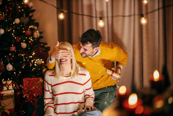 若い男はクリスマスプレゼントで彼のガールフレンドが居心地の良い家でクリスマスと新年のイブを祝っているときに驚いています. - 写真・画像