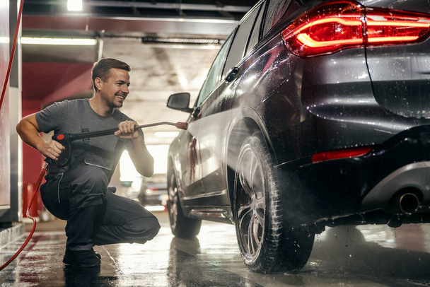 Šťastný umývač aut se krčí a myje auto pneumatiky s vysokotlakou vodní pistolí na stanici. - Fotografie, Obrázek