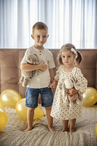 Retrato de hermanos felices parados en una cama rodeados de globos mientras sostienen sus manos y abrazan juguetes. - Foto, Imagen