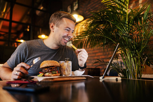 Un hombre feliz está comiendo su comida en el restaurante mientras tiene una llamada de conferencia con la familia en la tableta. - Foto, imagen