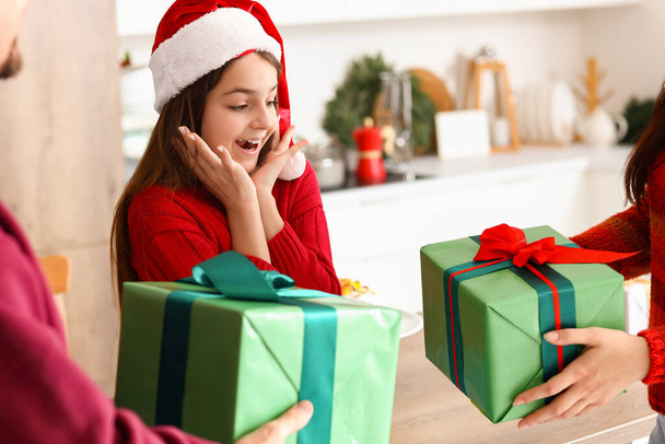 Удивлённая маленькая девочка получает рождественские подарки от родителей на кухне - Фото, изображение