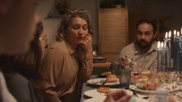 Média foto de preocupante mulher judia de meia-idade com cabelo encaracolado sentado à mesa durante a celebração Hanukkah família, falando com seus filhos adolescentes, enquanto o marido em kippah comer e ouvir - Filmagem, Vídeo