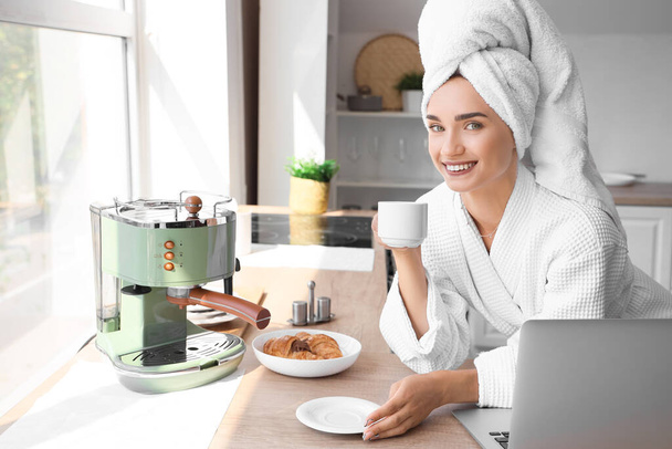 Schöne junge Frau im Bademantel trinkt Kaffee und benutzt Laptop in der Küche - Foto, Bild