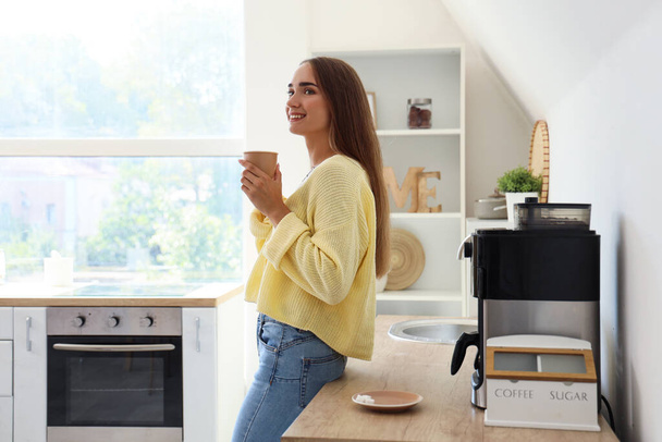 Beautiful young woman drinking hot espresso near modern coffee machine in kitchen - Photo, Image