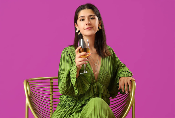 Молодая женщина с бокалом вина сидит в кресле на фиолетовом фоне - Фото, изображение