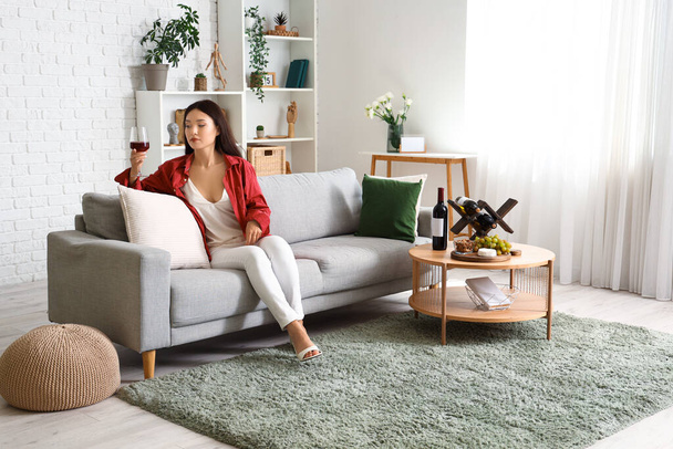 Mulher asiática bonita jovem com copo de vinho e lanches sentado no sofá na sala de estar - Foto, Imagem