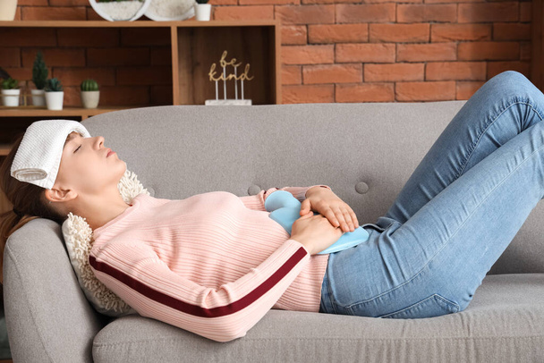 Kranke junge Frau mit Wärmflasche leidet unter Bauchschmerzen auf dem heimischen Sofa - Foto, Bild