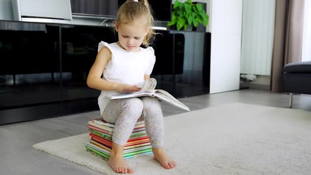 La niña está sentada en una pila de libros para niños y hojeando un libro con cuentos de hadas. Imágenes de alta calidad 4k - Metraje, vídeo