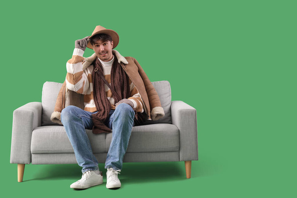 Young man in winter clothes sitting on grey sofa against green background - Photo, Image