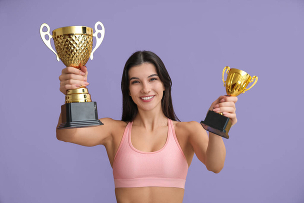 Mujer joven deportiva con copas de oro sobre fondo lila - Foto, imagen