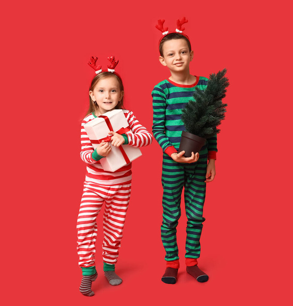 Cute little children in pajamas with Christmas gift and tree on red background - Photo, Image