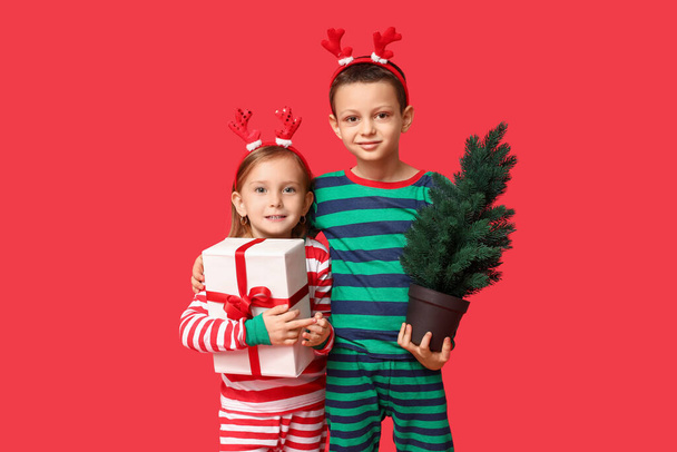 Милі маленькі діти в піжамі з різдвяним подарунком і деревом на червоному тлі - Фото, зображення