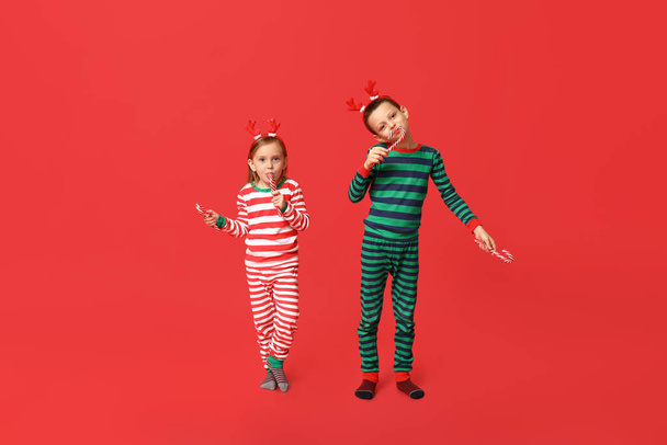 Lindos niños pequeños en pijama con bastones de caramelo sobre fondo rojo - Foto, Imagen