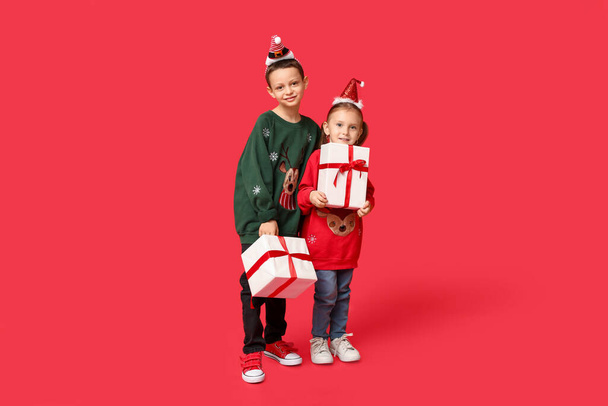 Cute little children in Christmas sweaters with gifts on red background - Photo, Image